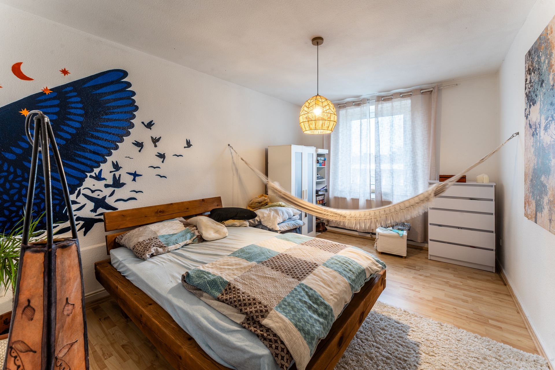 Helles Schlafzimmer mit Bett, Hängematte, Kleiderschrank und Wandbild mit blauen Vögeln.