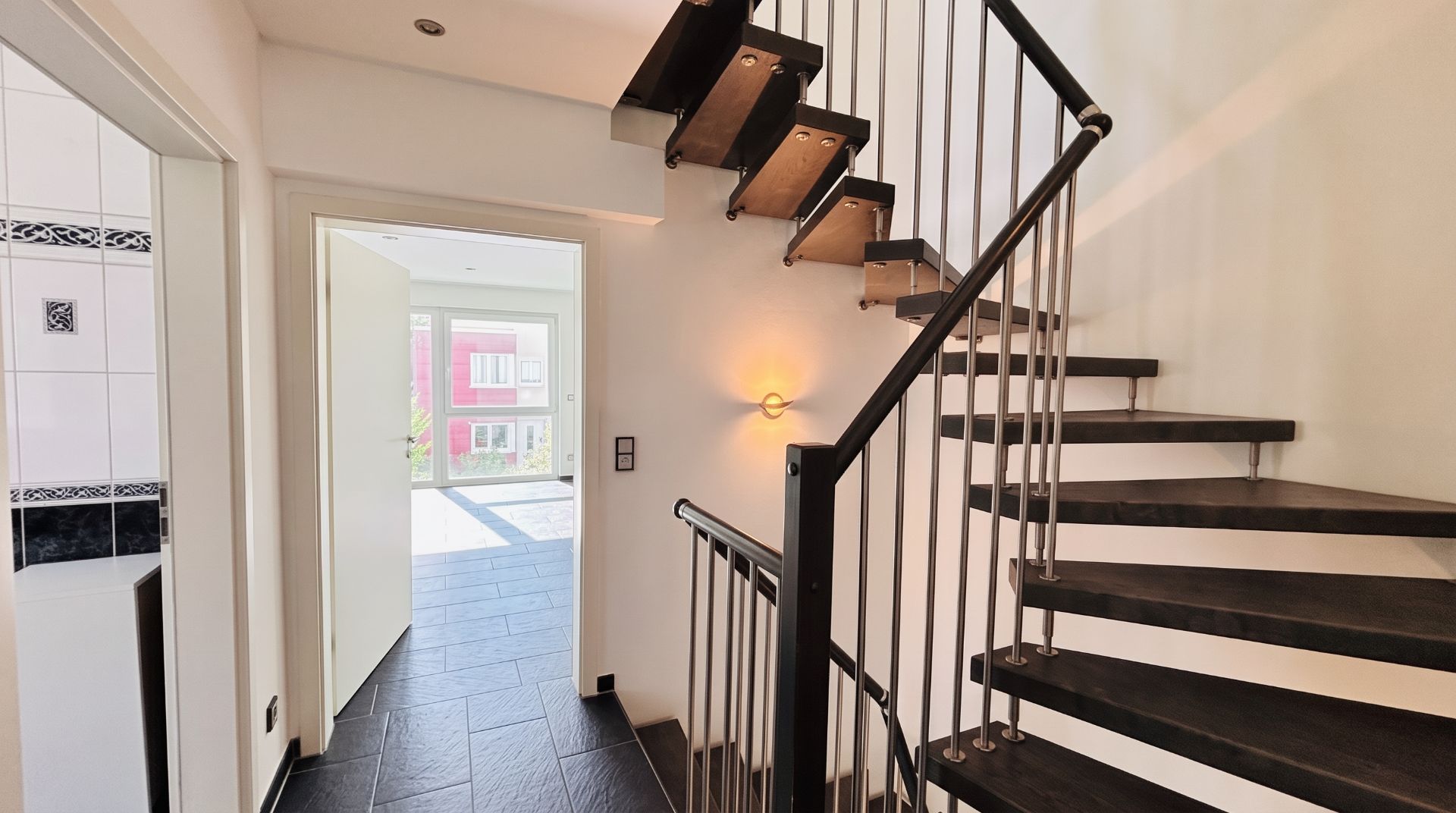 Heller Flur mit moderner, offener Treppe und Blick in einen Wohnraum mit großen Fenstern.