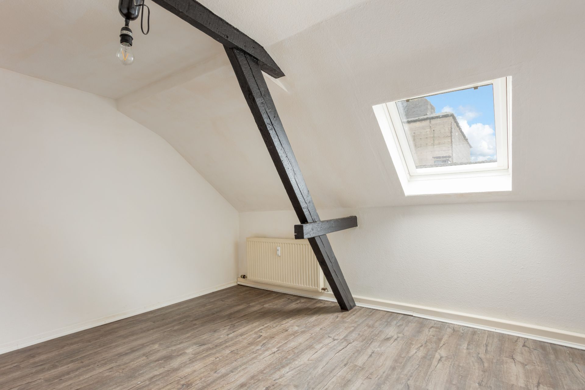 Helles Dachzimmer mit Dachfenster, sichtbarem Balken, Heizkörper und Holzfußboden.