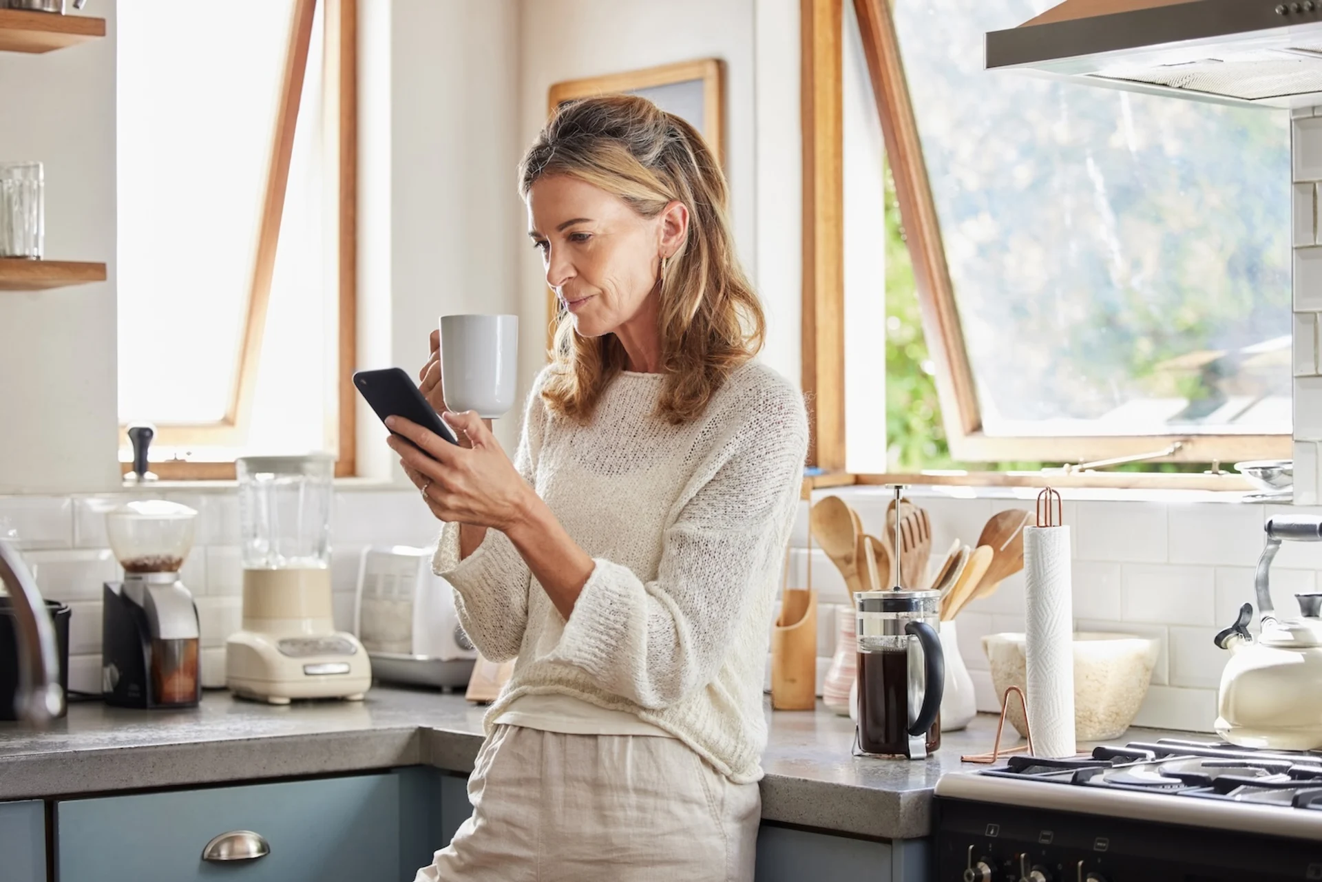 Frau steht in heller Küche, hält Tasse und Smartphone, Kaffeemaschine und Küchenutensilien sichtbar.