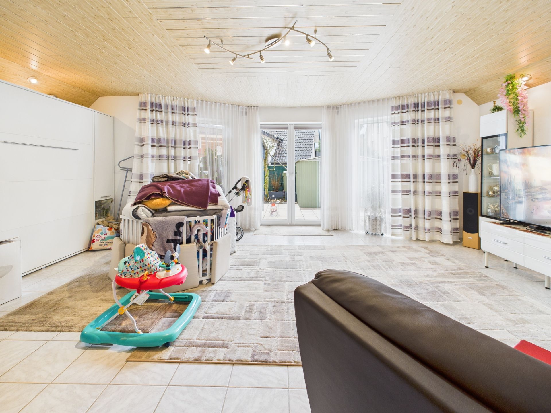 Helles Wohnzimmer mit Kinderbett, Spielzeug und großen Fenstern mit Vorhängen.
