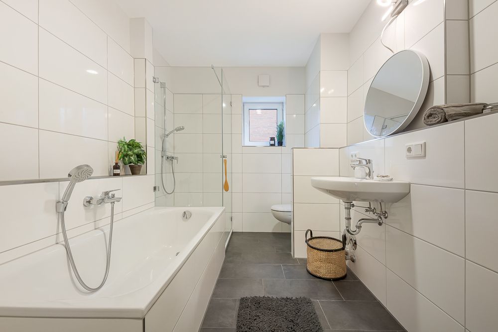 Modernes Badezimmer mit Badewanne, Dusche, Waschbecken, rundem Spiegel und Fenster.