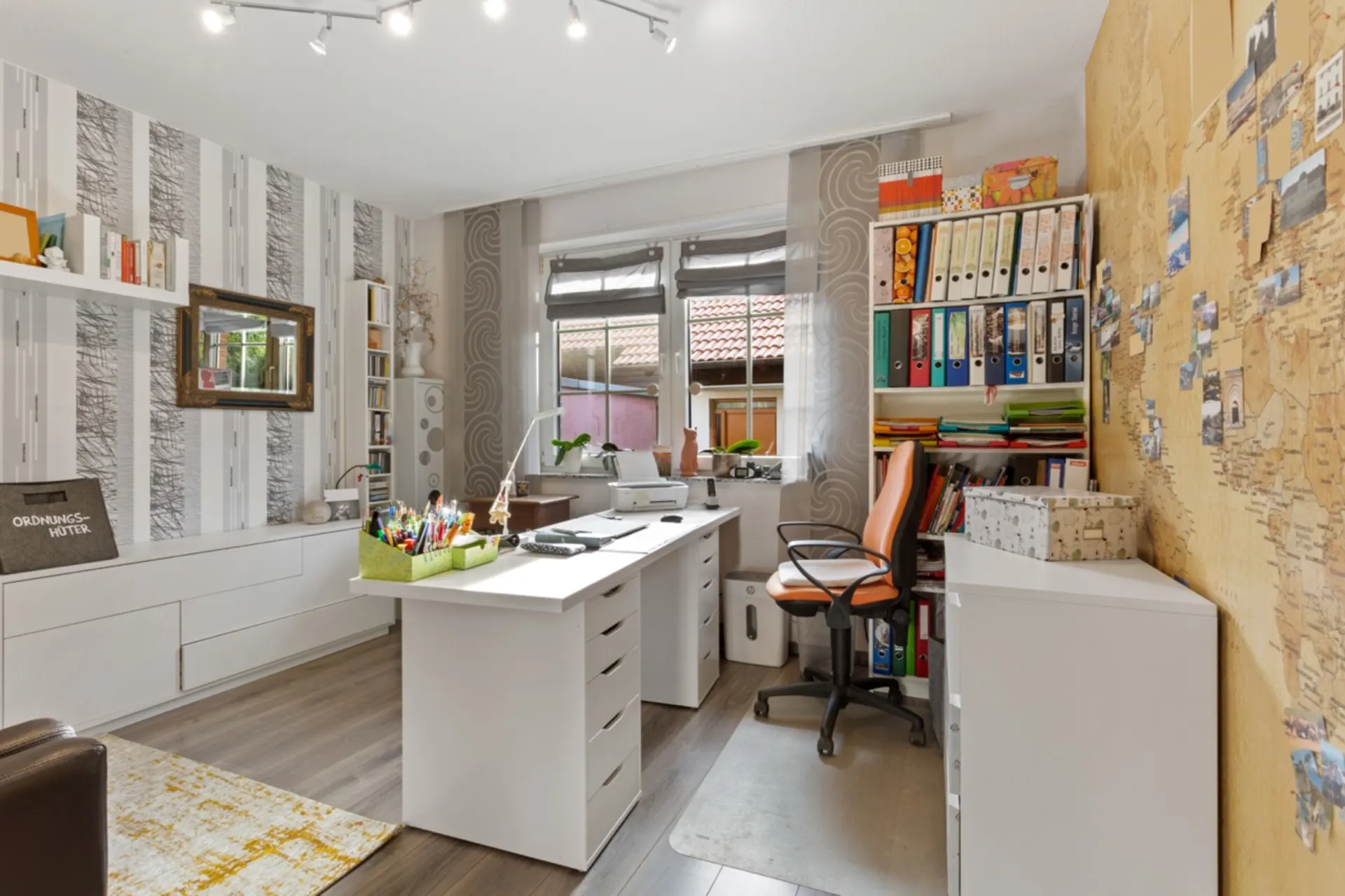 Helles Arbeitszimmer mit Schreibtisch, Bürostuhl, Regalen und Weltkarten-Tapete an der Wand.