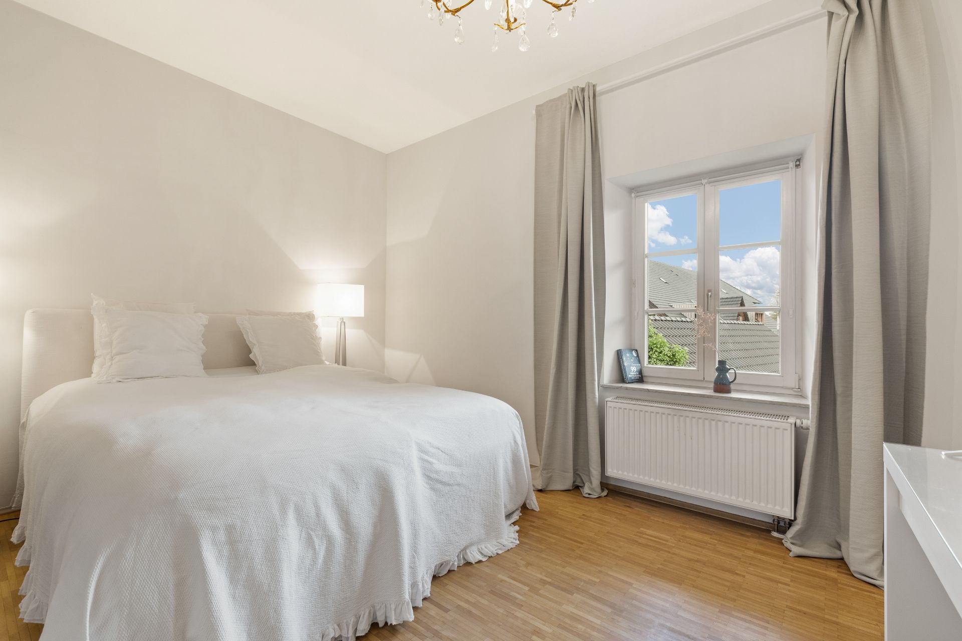 Helles Schlafzimmer mit Doppelbett, Nachttischlampe und Fenster mit grauen Vorhängen.
