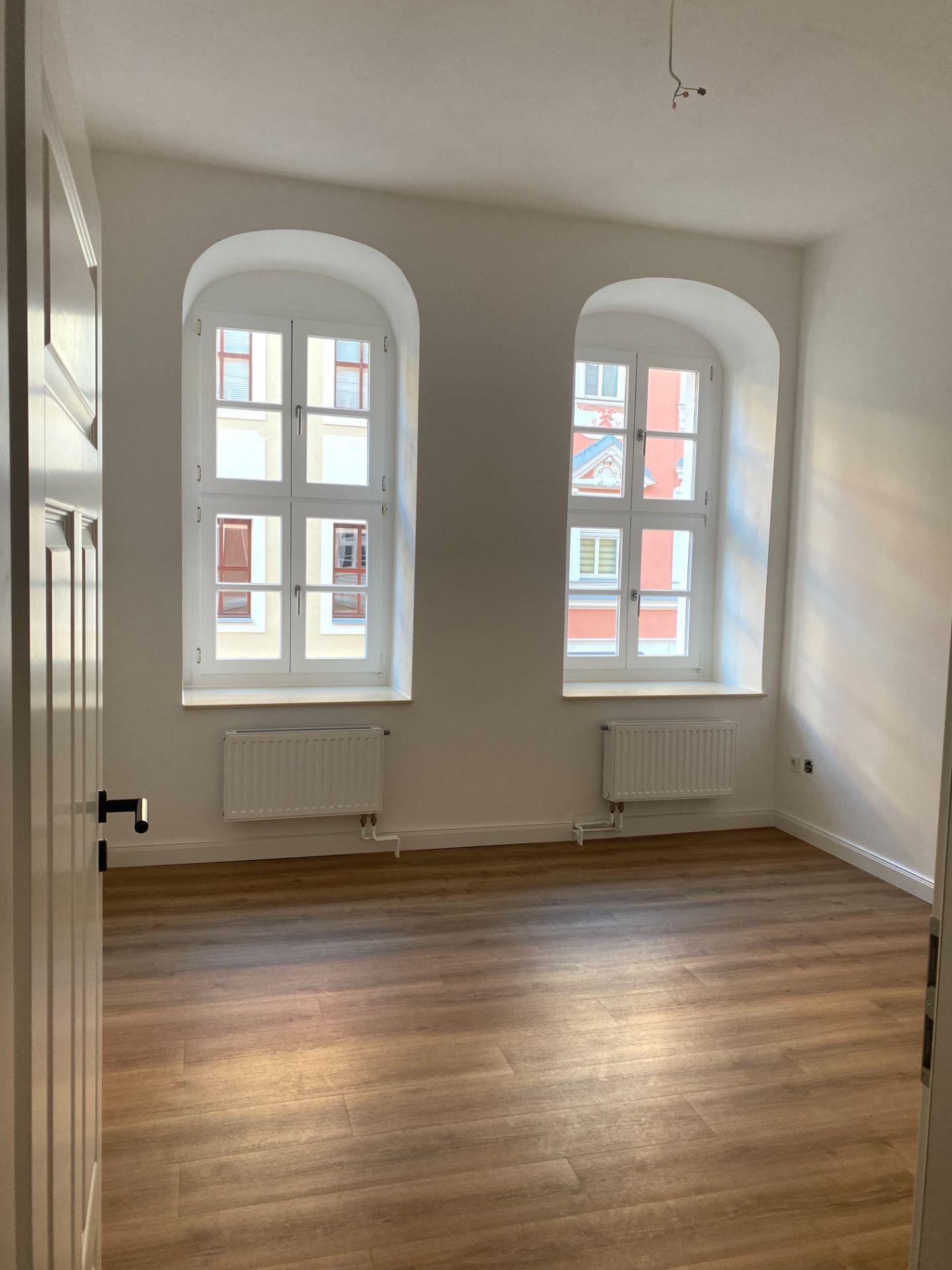 Heller Raum mit zwei großen Fenstern, weißer Wand und Holzfußboden.