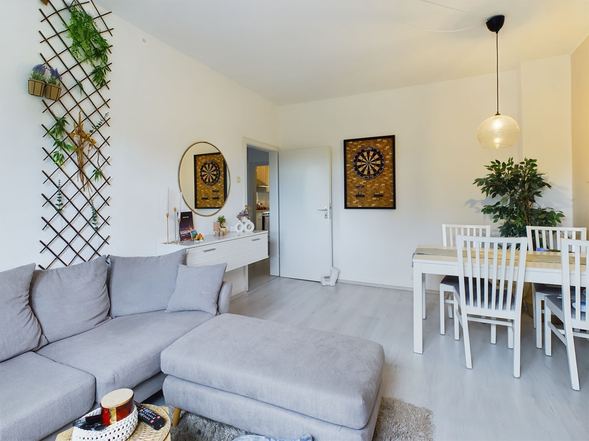 Wohnzimmer mit grauem Sofa, Esstisch mit weißen Stühlen, Wanddeko und Pflanze.