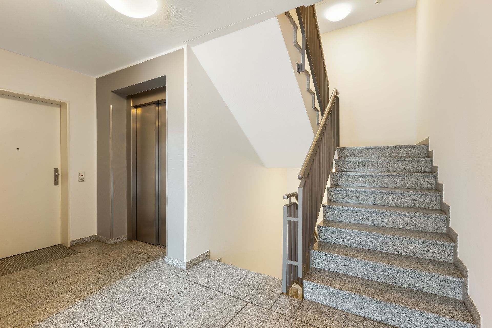 Treppenhaus mit Aufzug und Wohnungstür, Wände und Boden in neutralen Farben.