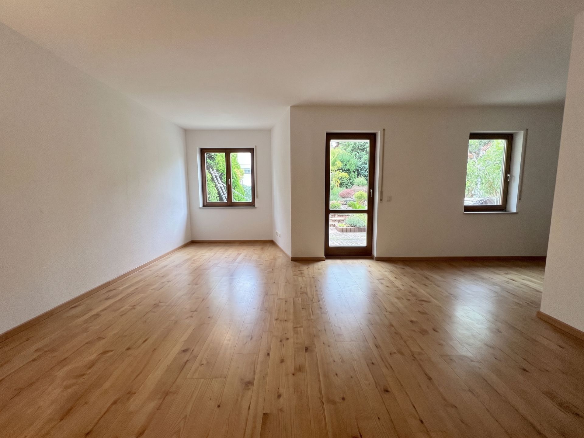 Helles, leeres Zimmer mit Holzfußboden, zwei Fenstern und einer Tür zum Garten.