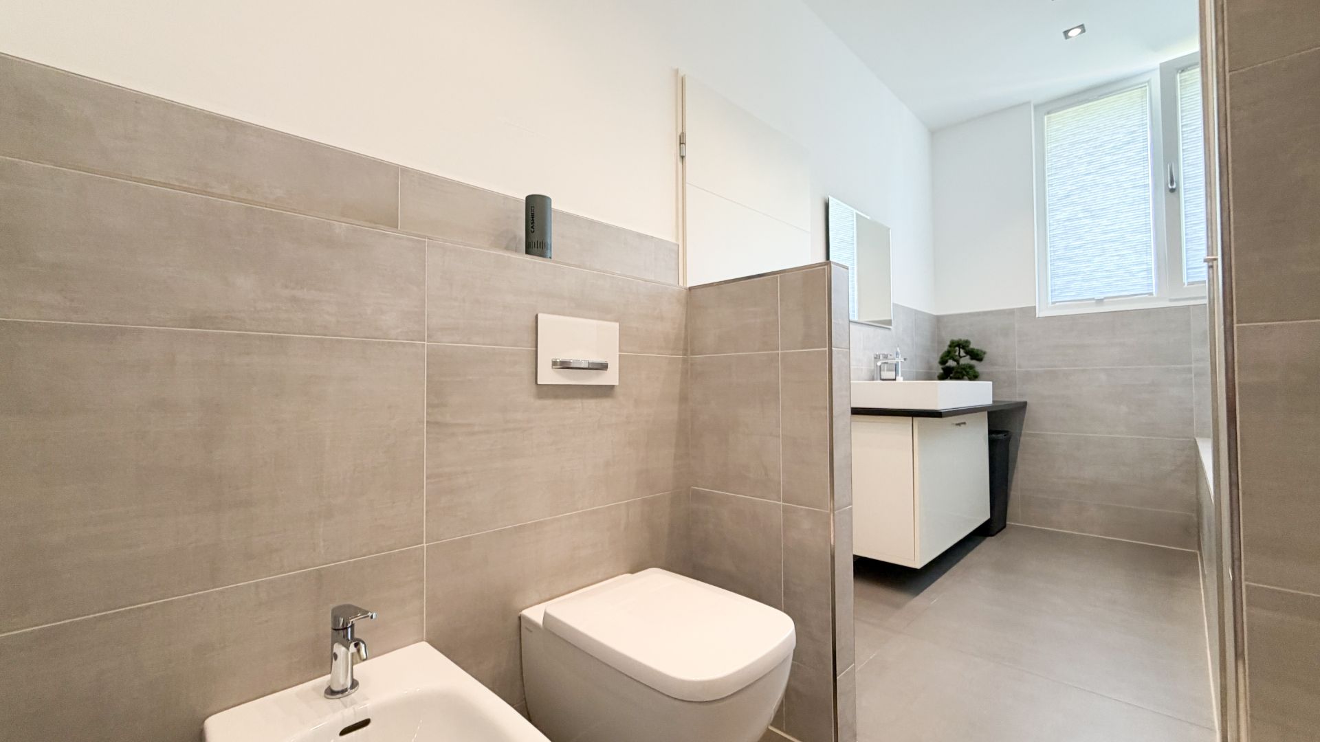 Modernes Bad mit Toilette, Bidet, Waschbecken und großem Fenster, helle Fliesen.