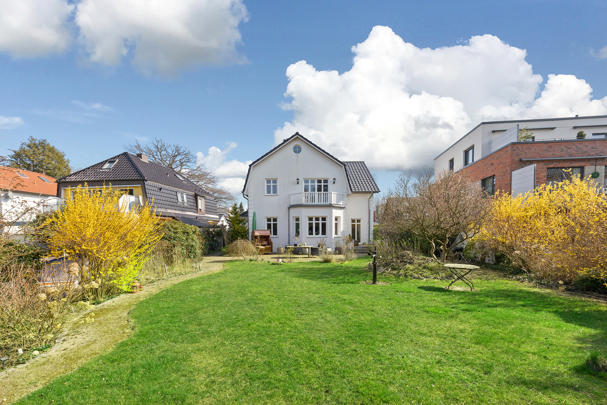 Weißes Wohnhaus mit Balkon und großem Garten, umgeben von Sträuchern und Nachbarhäusern.