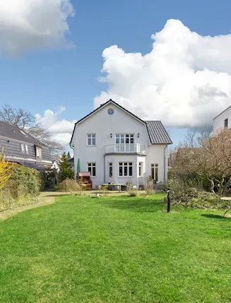 Weißes Wohnhaus mit Balkon und großem Garten, umgeben von Sträuchern und Nachbarhäusern.