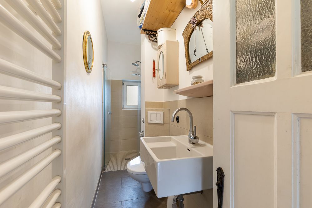 Schmales Badezimmer mit Waschbecken, Spiegel, Toilette und Dusche am Fenster.