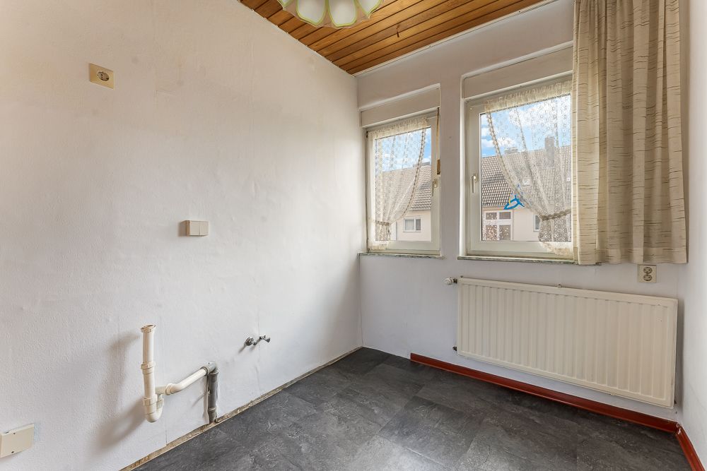 Leerer Raum mit zwei Fenstern, Heizung und sichtbaren Rohren an der Wand.