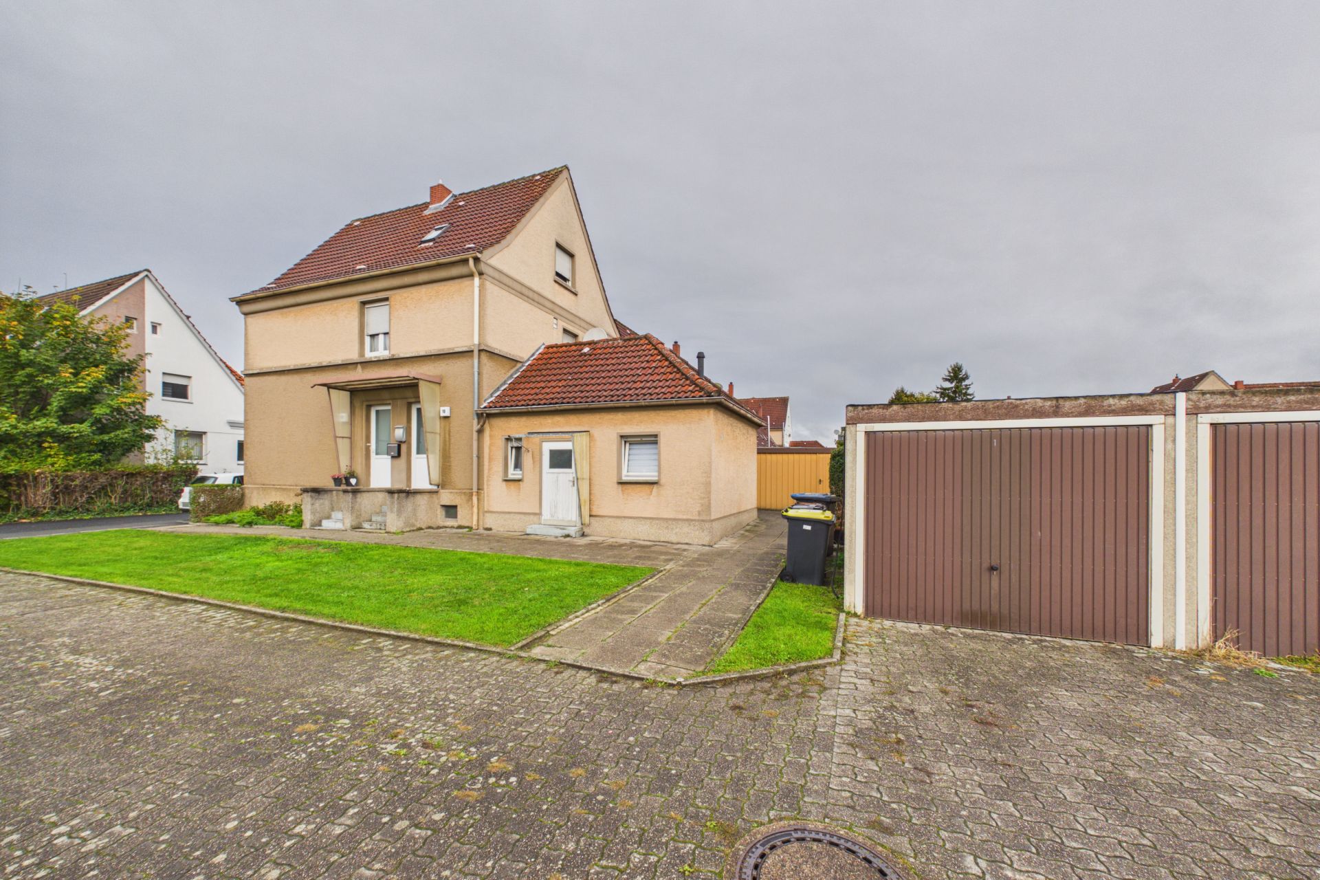 Beiges Wohnhaus mit rotem Dach, Vorgarten und gepflastertem Weg, Garage rechts im Bild.