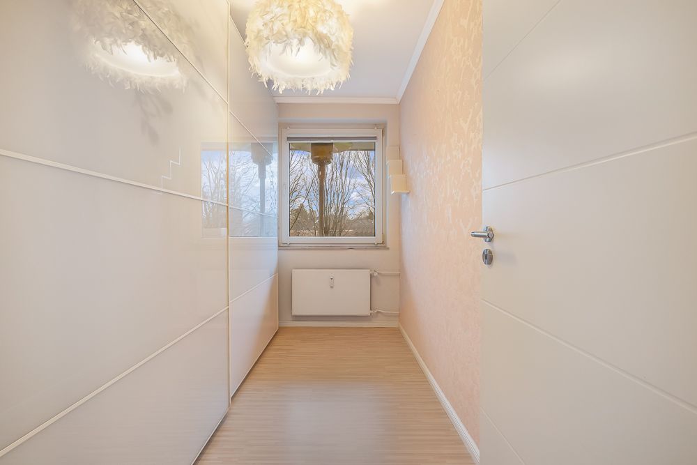 Schmaler, heller Raum mit weißer Tür, Kleiderschrank, Fenster und modernem Deckenlicht.