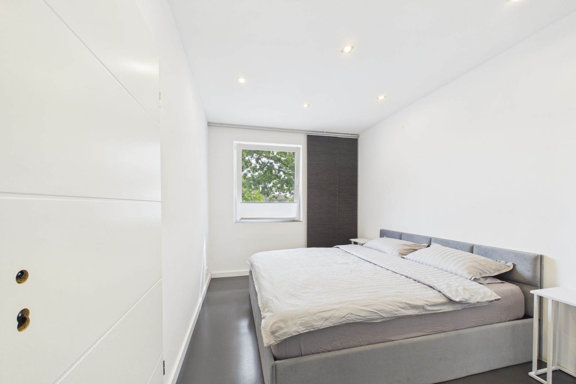 Helles Schlafzimmer mit Doppelbett, Nachttisch und Fenster mit Blick auf Bäume.