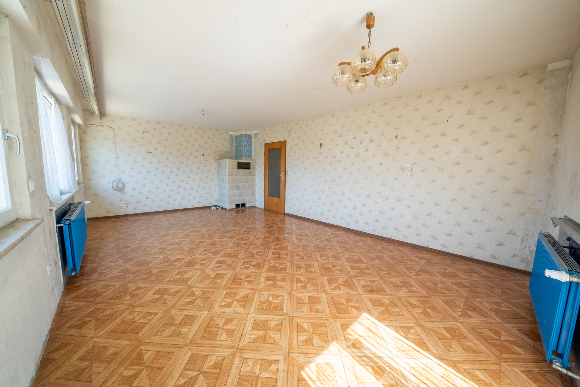 Leeres Zimmer mit Holzfußboden, Tapeten, drei Fenstern, blauem Heizkörper und Deckenlampe.
