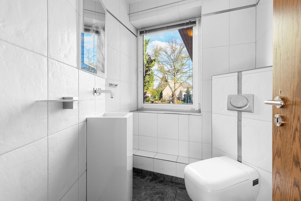 Modernes, weiß gefliestes WC mit kleinem Waschbecken und großem Fenster.
