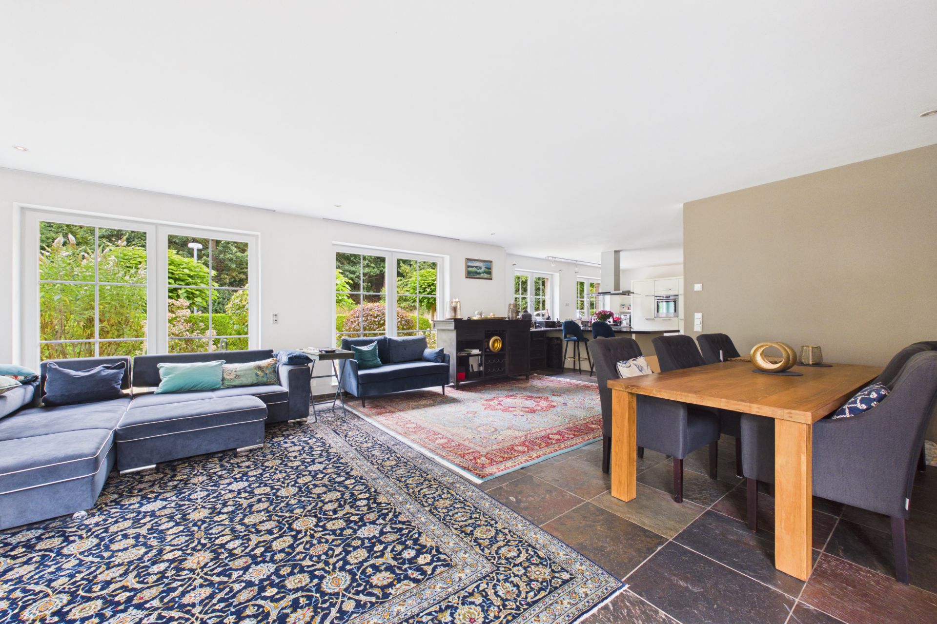 Helles Wohnzimmer mit großen Fenstern, Sofas, Teppichen und einem Esstisch mit Stühlen.