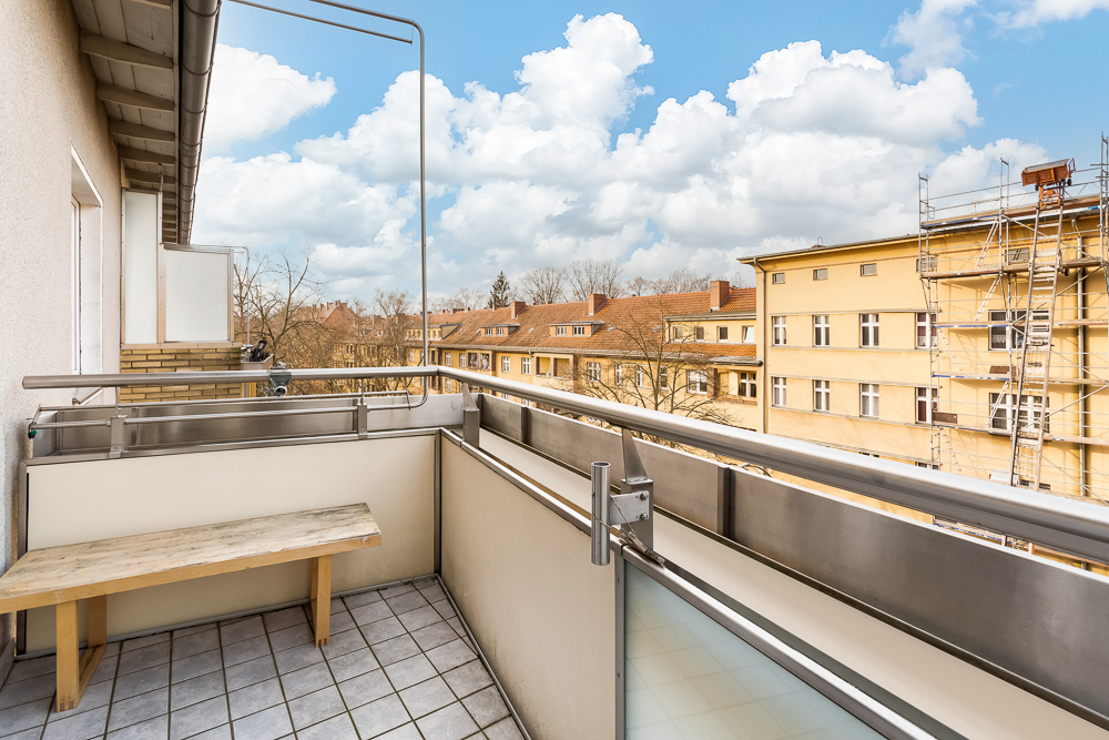 Balkon mit Holzbänkchen, Fliesenboden und Ausblick auf Nachbargebäude unter blauem Himmel.