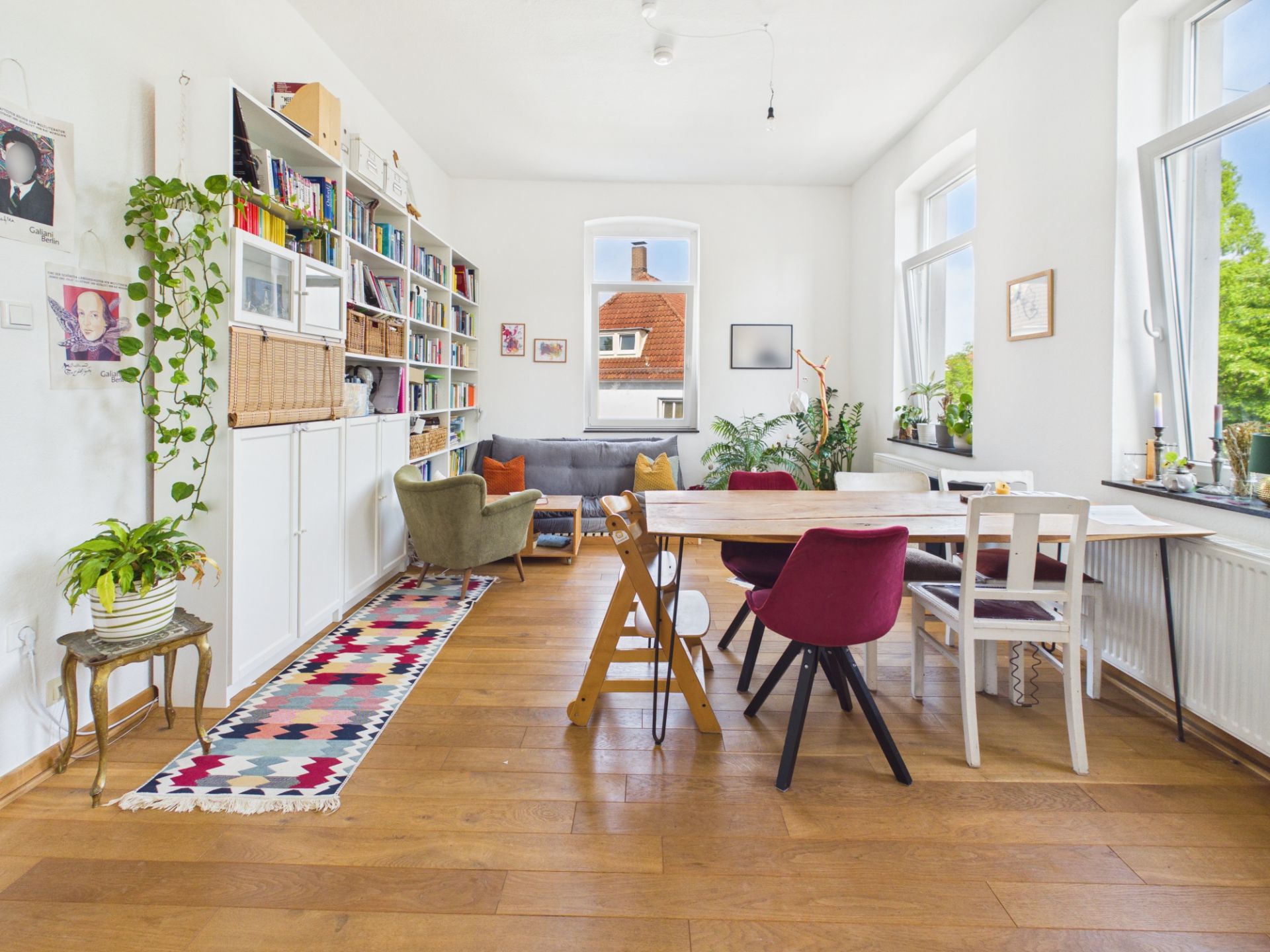 Helles Wohnzimmer mit Esstisch, bunten Stühlen, Regalwand, Sofa und großen Fenstern.