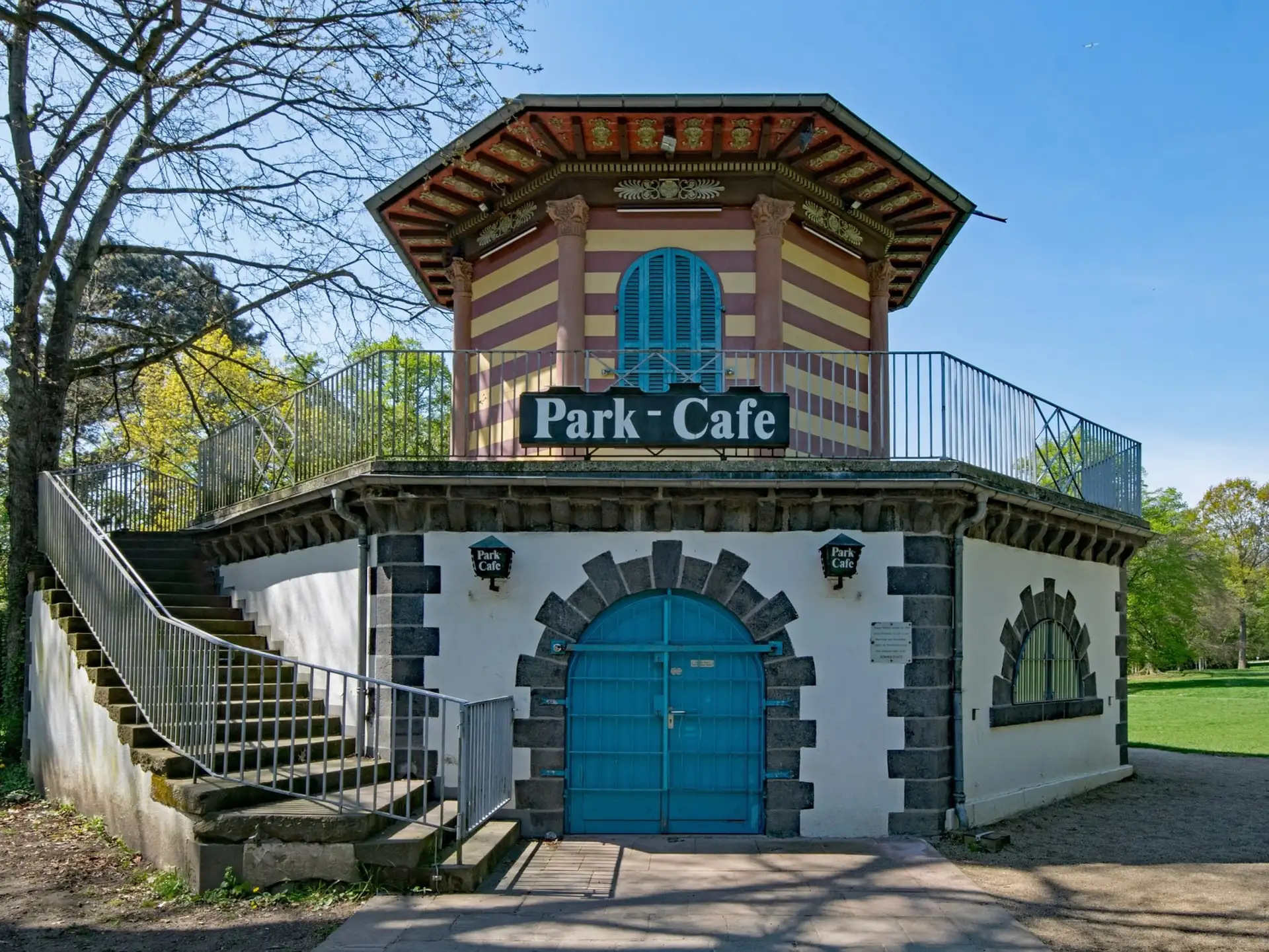 Zweistöckiges Gebäude mit blauen Türen und Fenstern, Schild 'Park Cafe', Treppe links, umgeben von Bäumen.