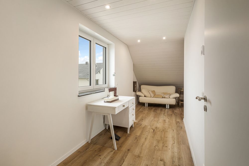Helles Zimmer mit Holzboden, Schreibtisch am Fenster und Sofa an der Wand.