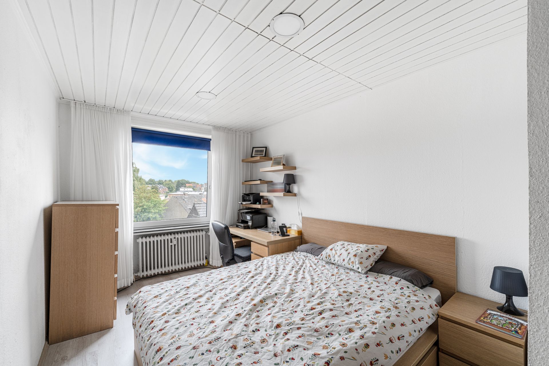 Helles Schlafzimmer mit großem Fenster, Bett, Schreibtisch und Kommode aus Holz.
