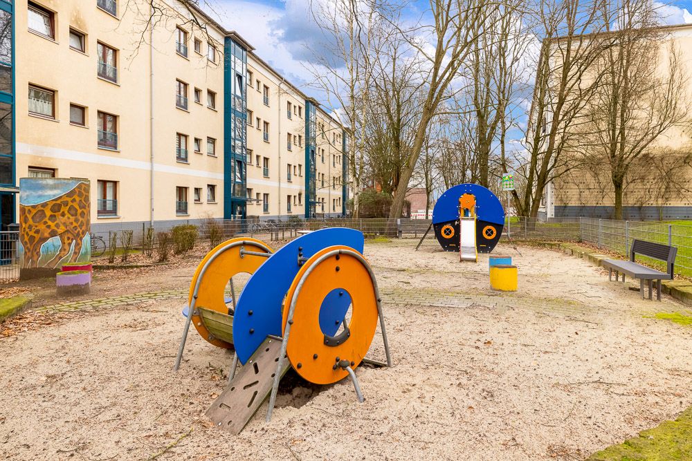 Spielplatz mit bunten Klettergeräten und Bänken vor Wohnhäusern, umgeben von Bäumen.