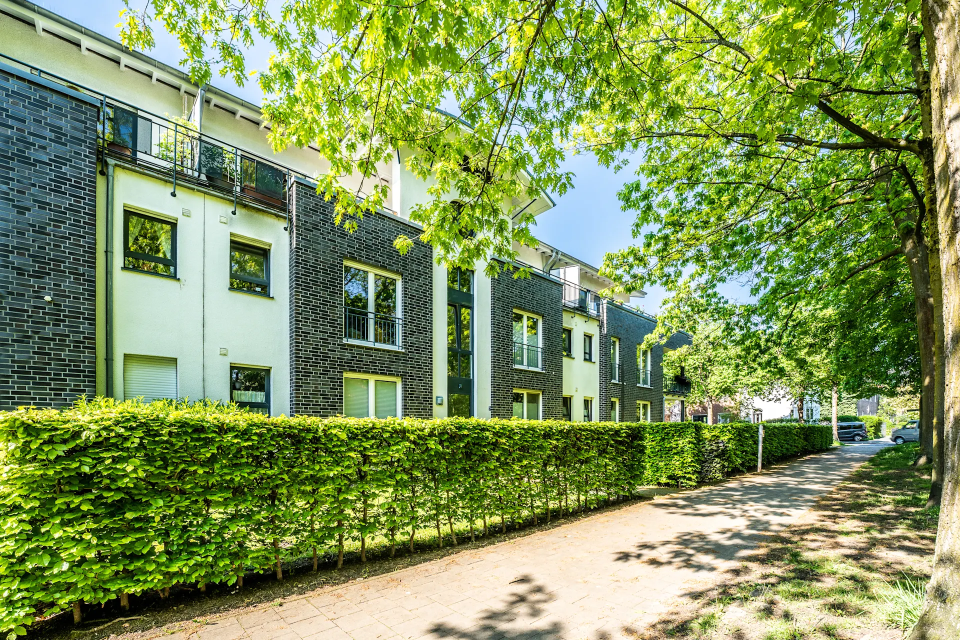 Modernes Wohngebäude mit Balkon, Hecke und Gehweg, umgeben von grünen Bäumen.