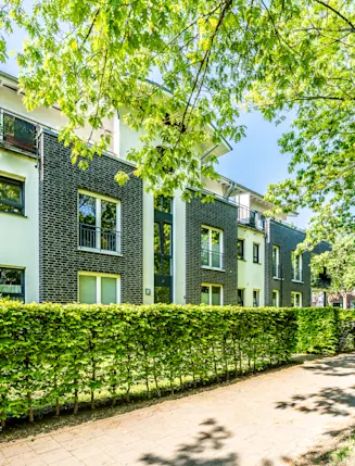 Modernes Wohngebäude mit Balkon, Hecke und Gehweg, umgeben von grünen Bäumen.