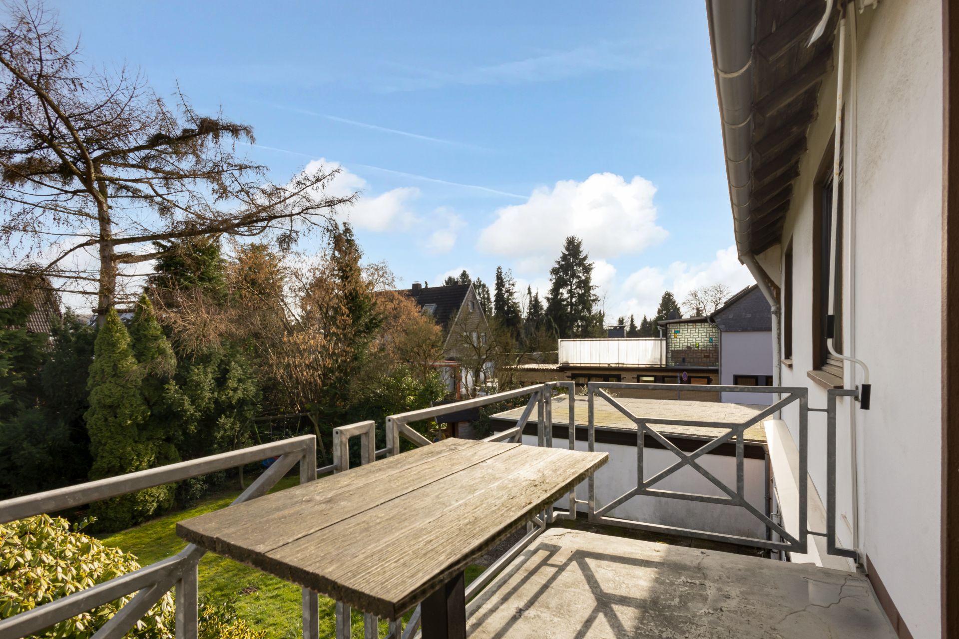 Balkon mit Holztisch und Geländer, Blick auf Garten und Bäume.