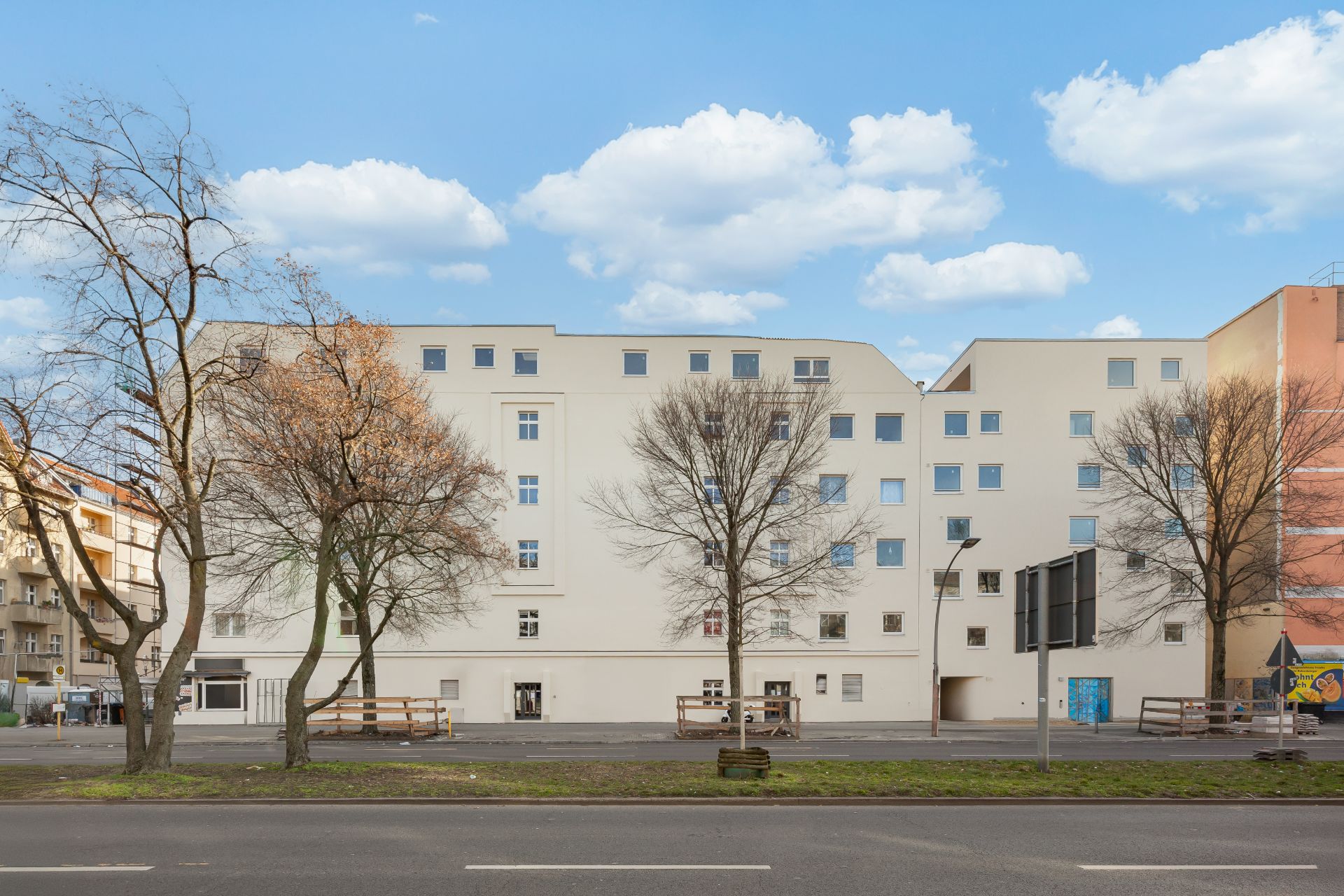 Mehrstöckiges Wohngebäude mit vielen Fenstern, Bäumen davor und einer Straße im Vordergrund.
