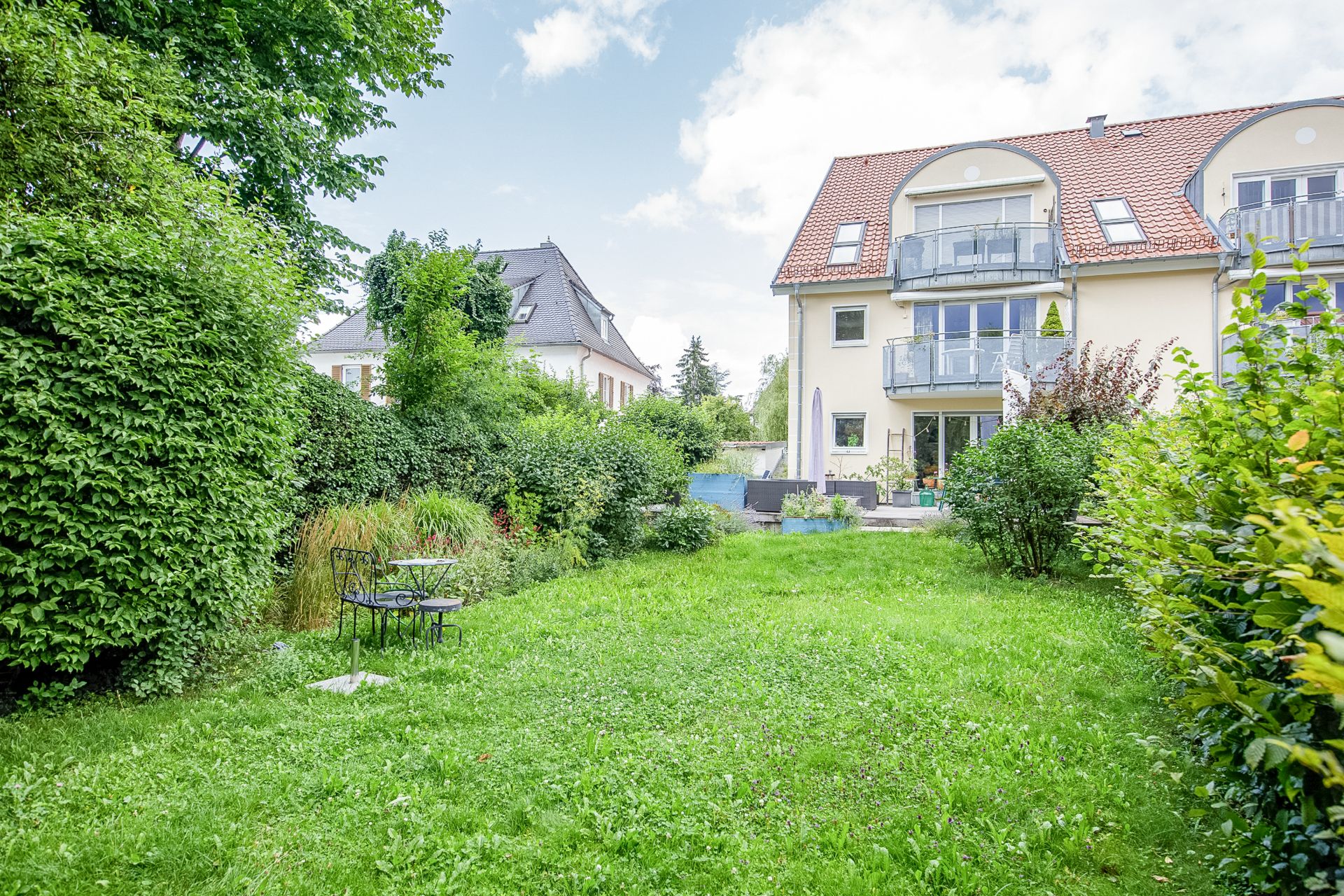 Grüner Garten mit Rasen, Gartenmöbeln und mehrstöckigem Wohnhaus im Hintergrund.