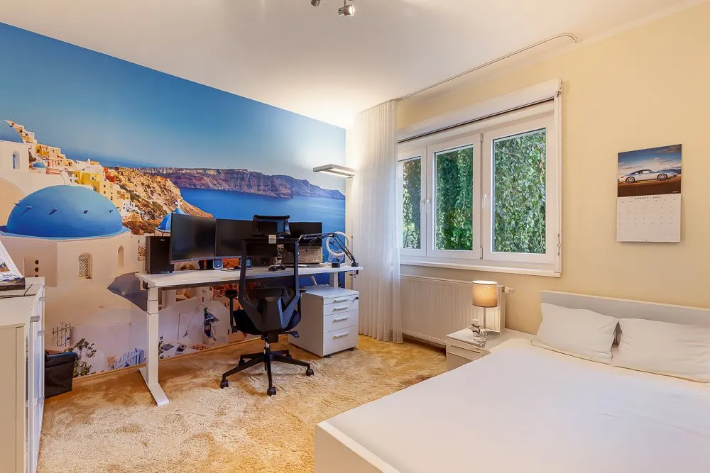 Helles Schlafzimmer mit Bett, Schreibtisch, Computer und einer Wand mit Santorini-Motiv.