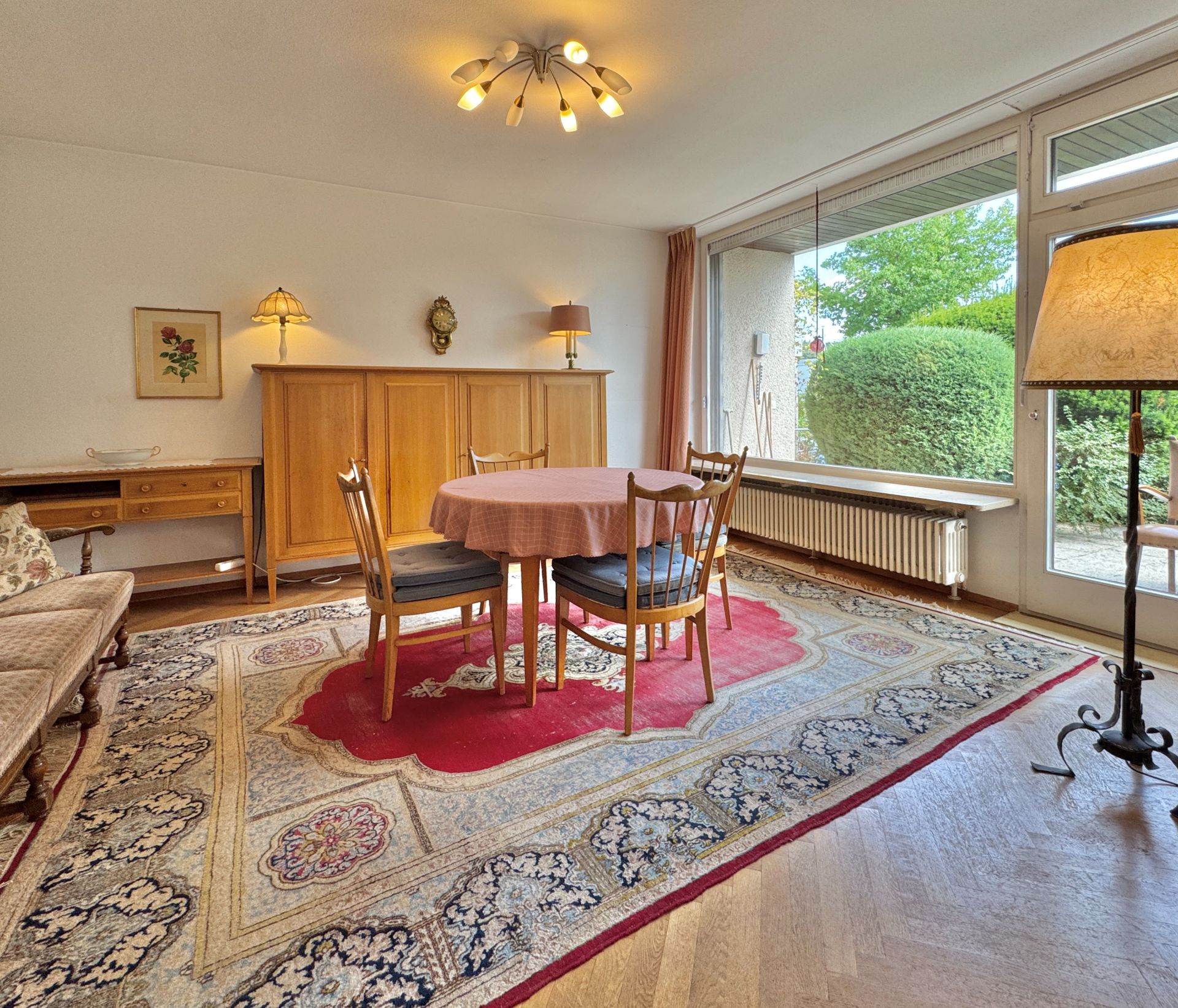 Wohnzimmer mit großem Teppich, rundem Tisch, vier Stühlen und großem Fenster zum Garten.