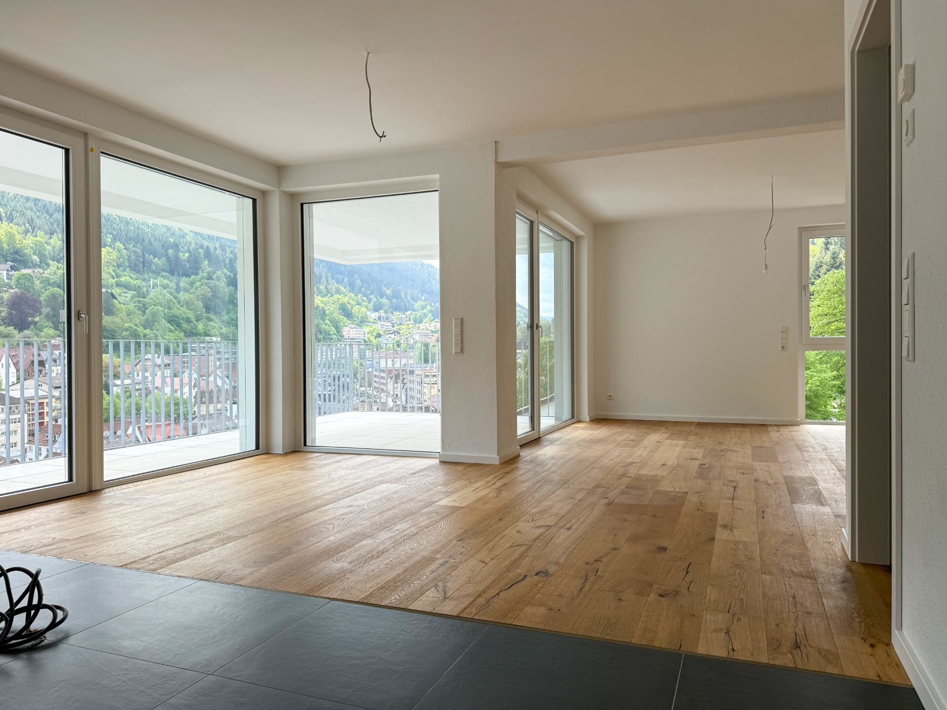 Helles, leeres Wohnzimmer mit Holzboden und großen Fenstern mit Blick auf grüne Hügel.