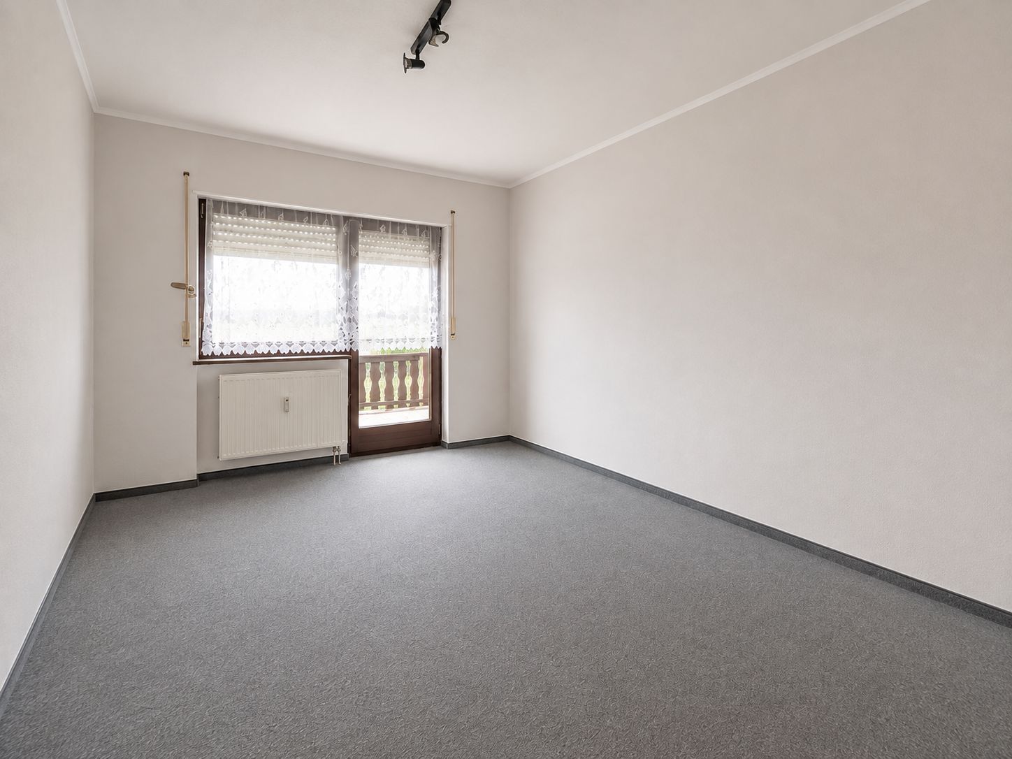 Leeres Zimmer mit grauem Teppichboden, großem Fenster und Tür zum Balkon.