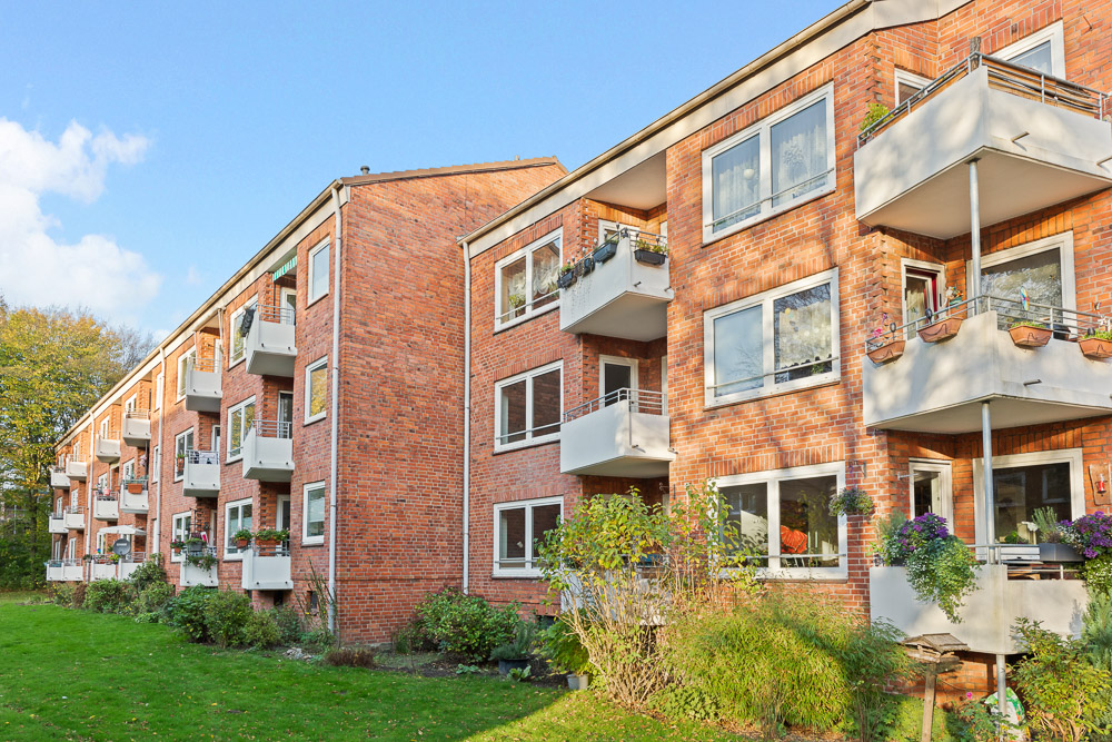 Rotes Mehrfamilienhaus mit weißen Balkonen und viel Grün im Vordergrund.