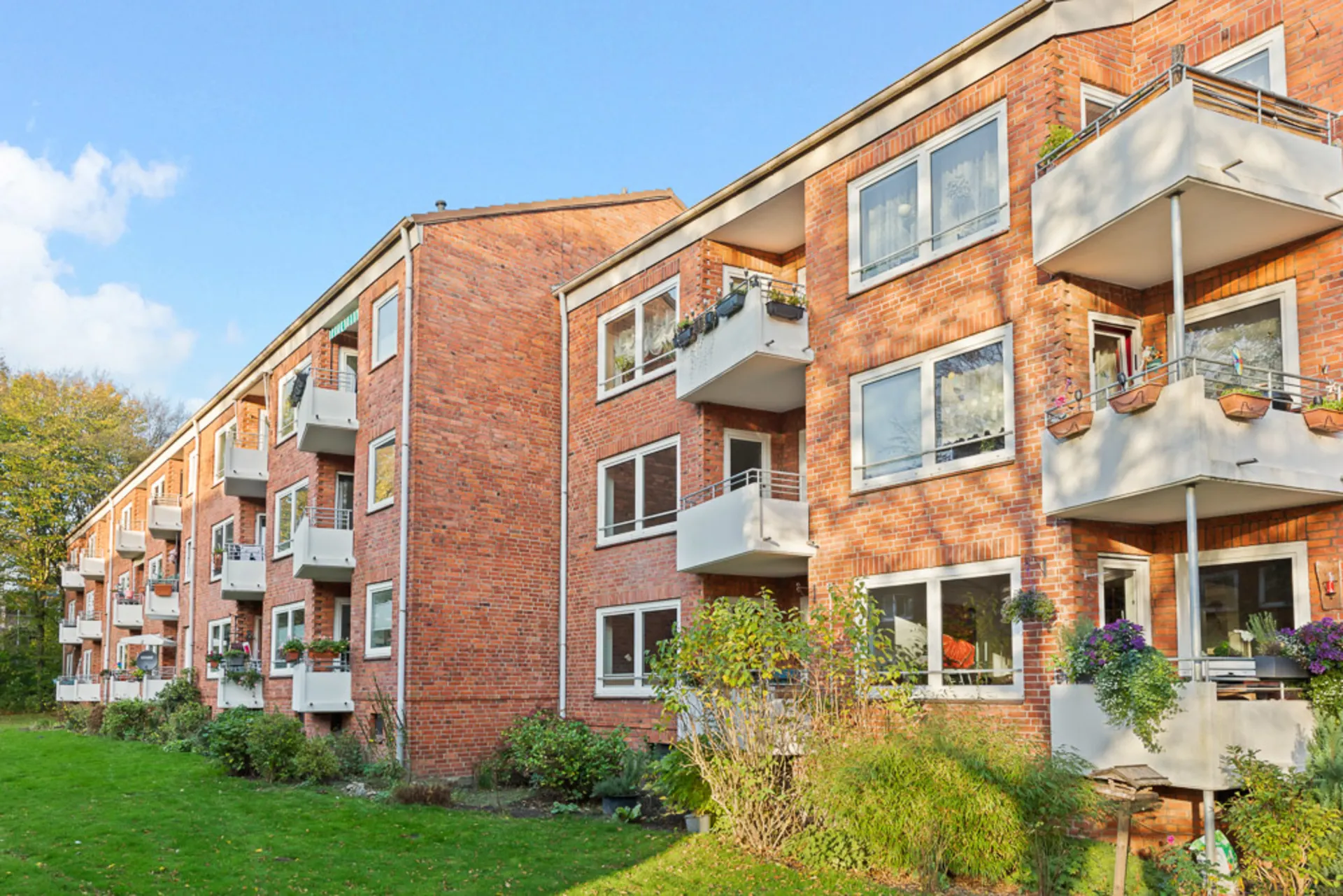 Rotes Mehrfamilienhaus mit weißen Balkonen und viel Grün im Vordergrund.
