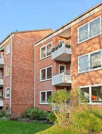 Rotes Mehrfamilienhaus mit weißen Balkonen und viel Grün im Vordergrund.