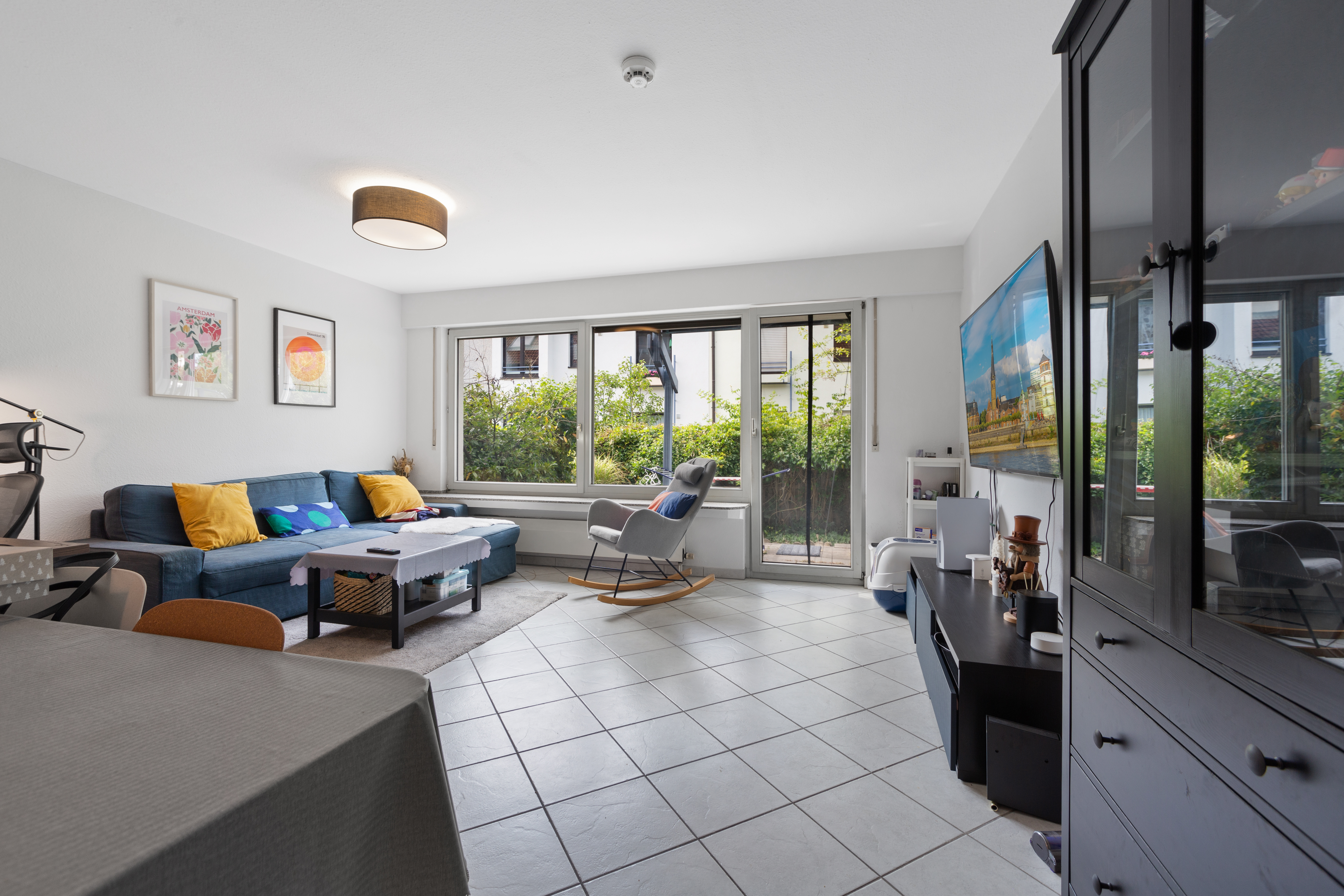 Helles Wohnzimmer mit Sofa, Couchtisch, Schaukelstuhl, großen Fenstern und Fernseher.