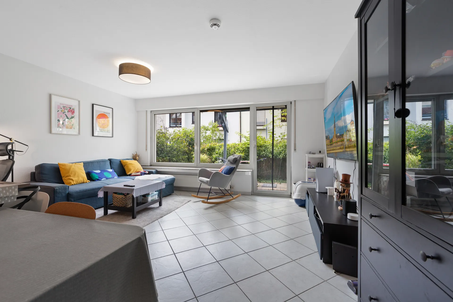 Helles Wohnzimmer mit Sofa, Couchtisch, Schaukelstuhl, großen Fenstern und Fernseher.
