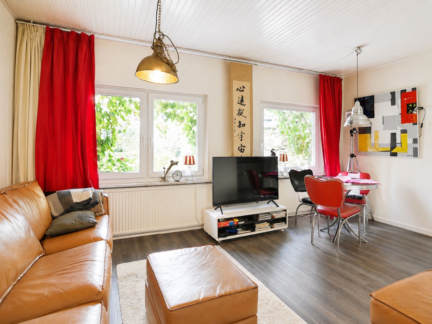 Wohnzimmer mit braunem Sofa, TV, roten Vorhängen und Esstisch mit roten Stühlen.