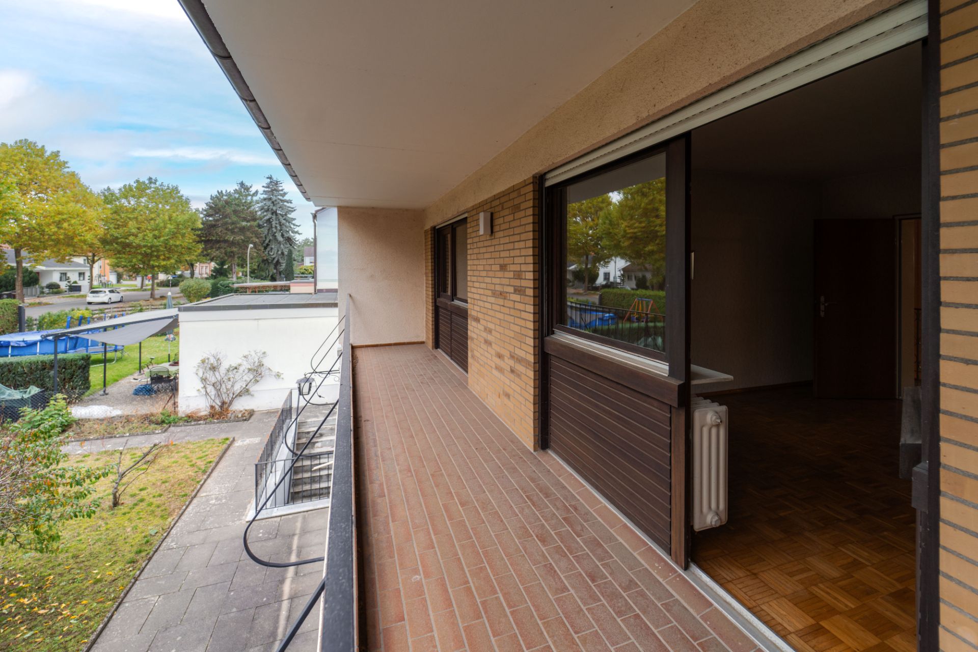 Langer Balkon mit Backsteinboden an einem Wohngebäude, Blick in den Garten und auf die Straße.