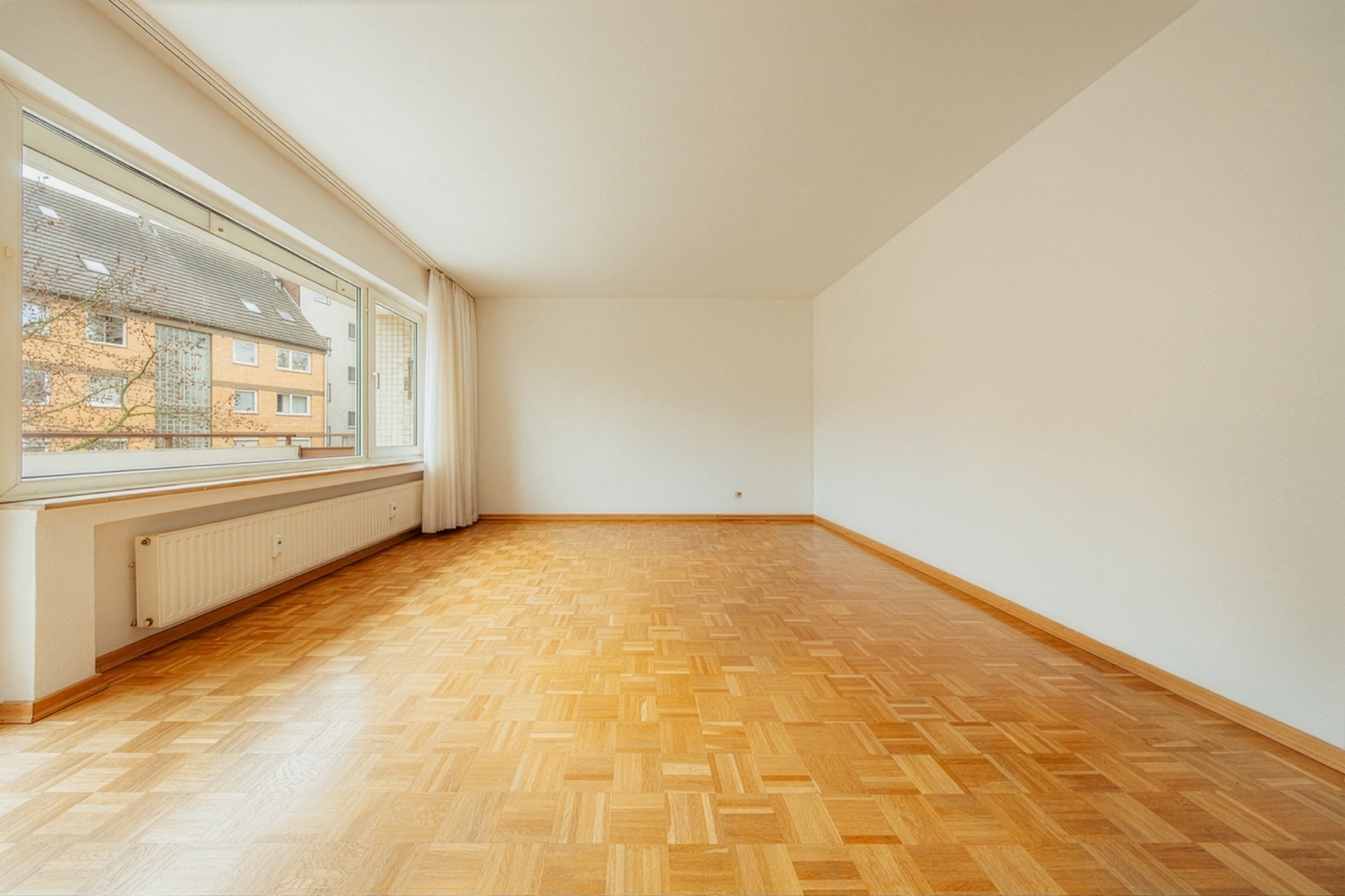 Leerer Raum mit Parkettboden, großem Fenster und weißen Wänden.