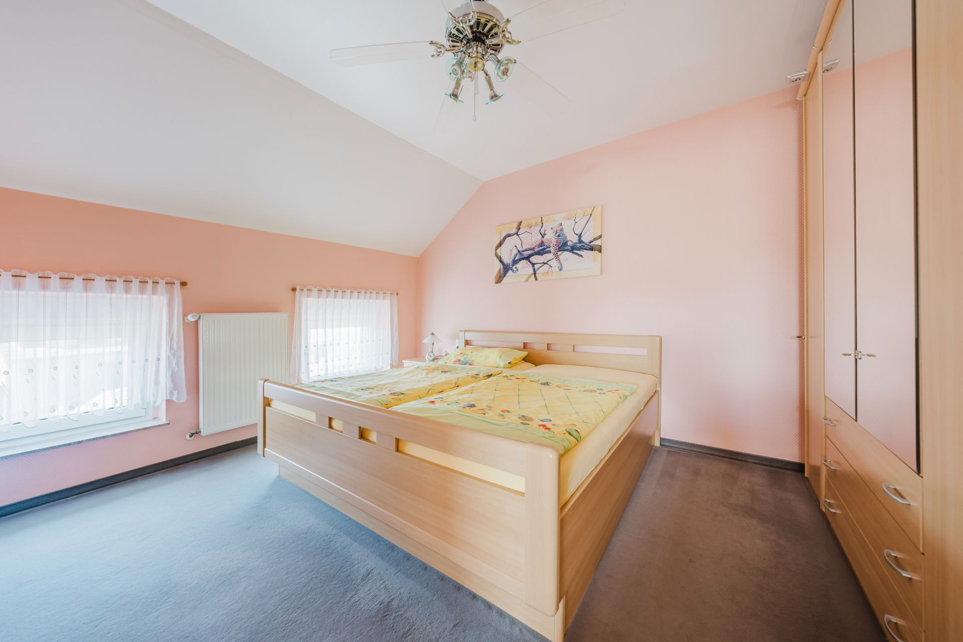 Helles Schlafzimmer mit Doppelbett, Kleiderschrank, zwei Fenstern und Deckenventilator.