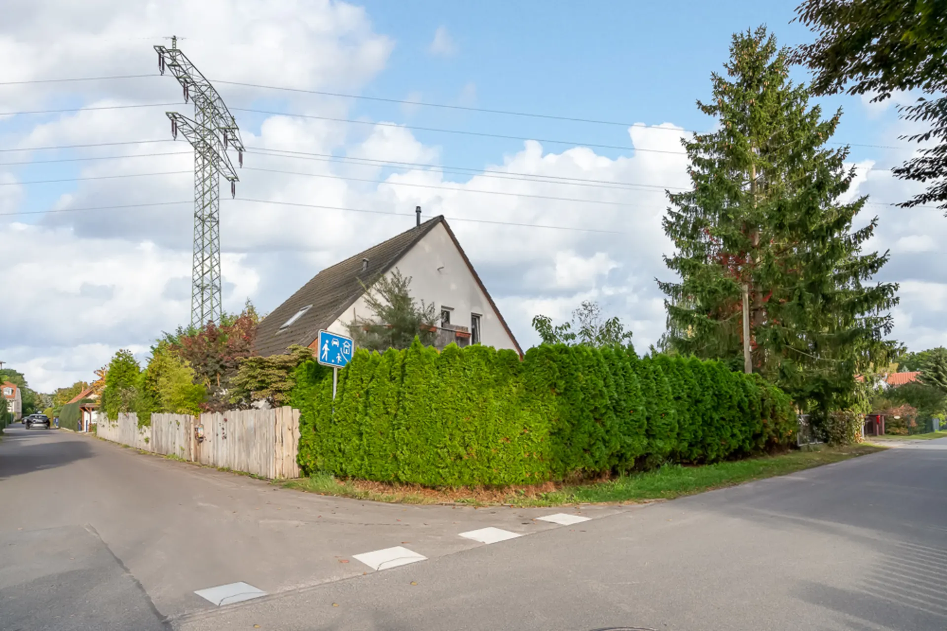 Einfamilienhaus mit schrägem Dach, hoher Hecke und Bäumen an einer Straßenecke.