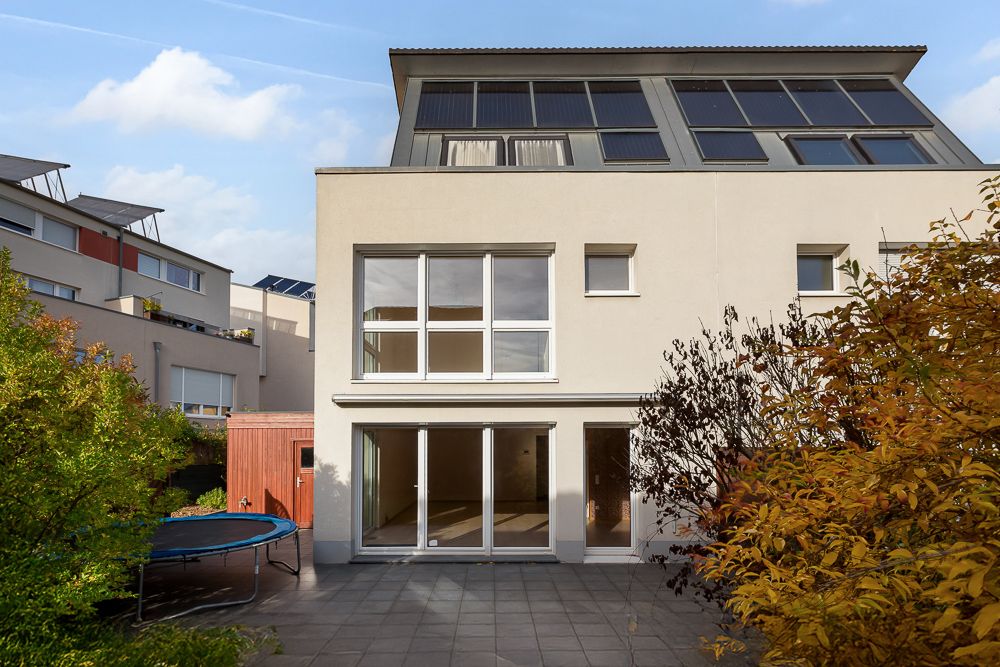 Moderne Rückseite eines Wohnhauses mit großen Fenstern, Terrasse und Trampolin im Garten.