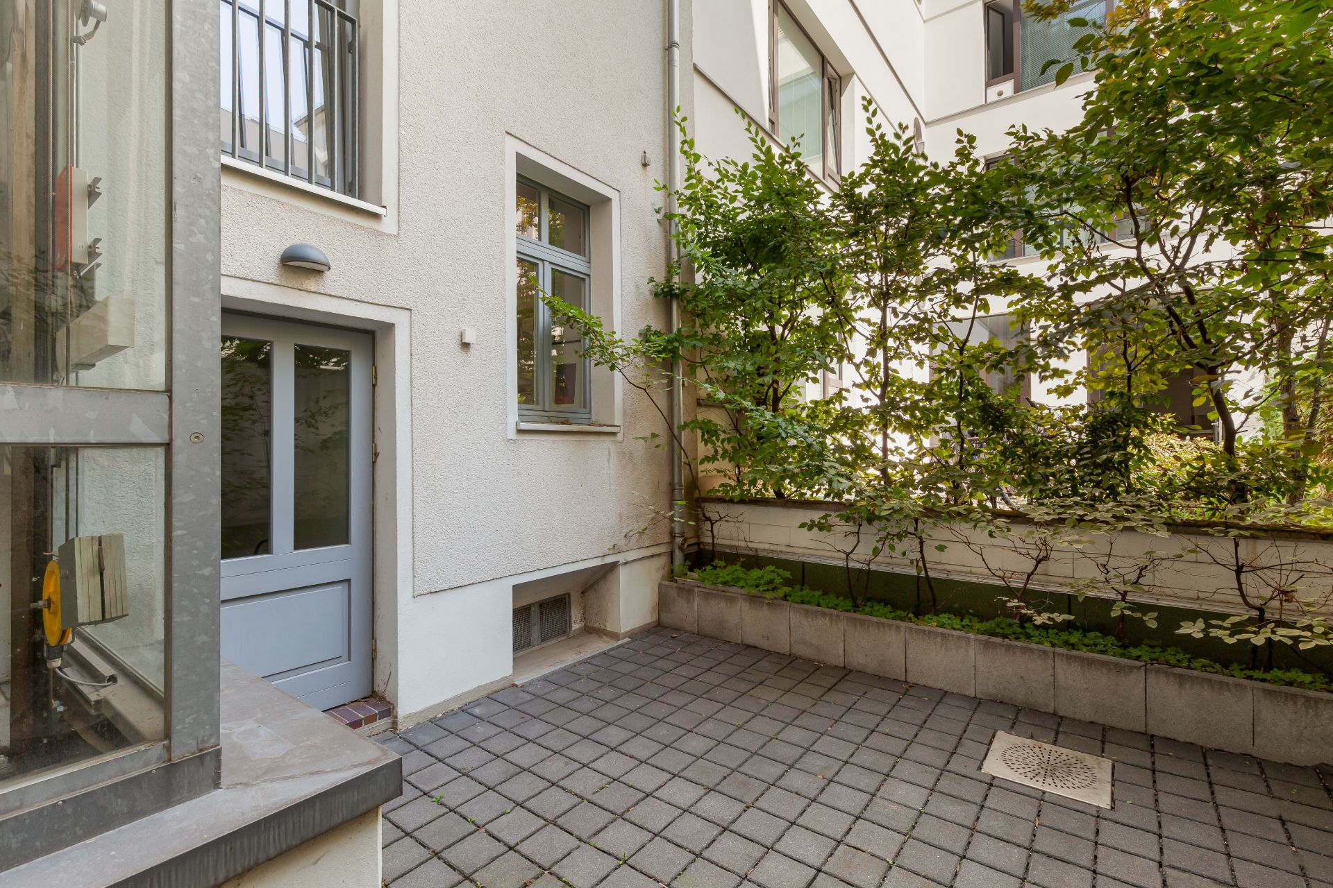 Gepflasterter Innenhof mit Tür, Fenster und bepflanztem Bereich an einer Wohnhauswand.